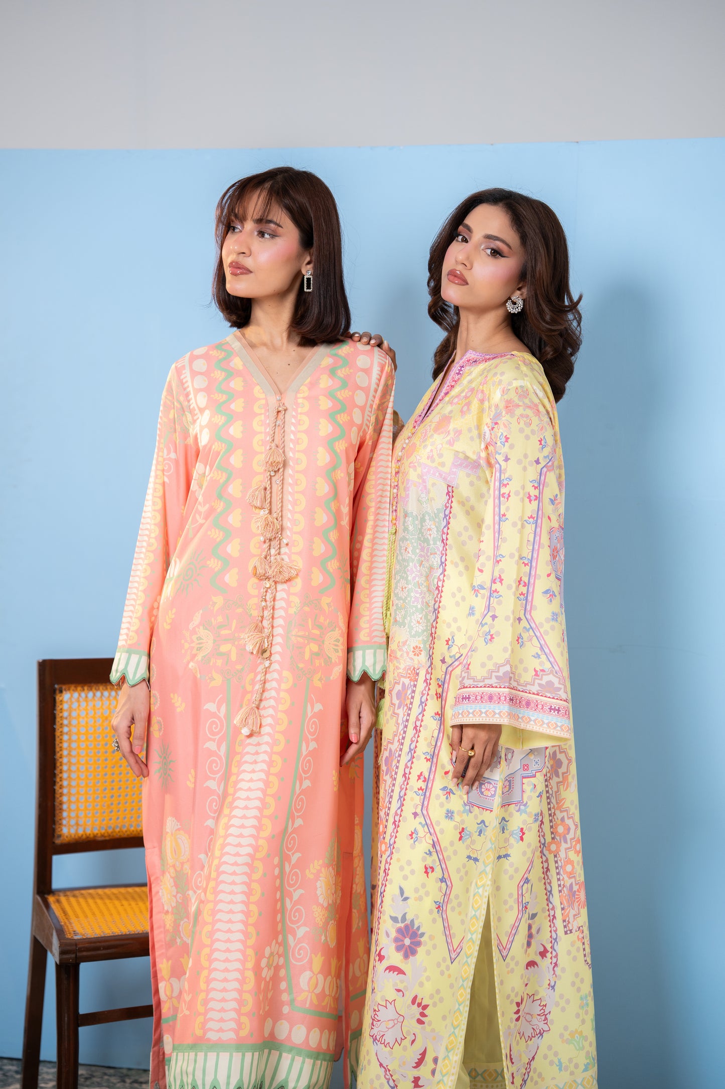 Two women wearing pastel-colored traditional outfits against a blue background SHAFAE
