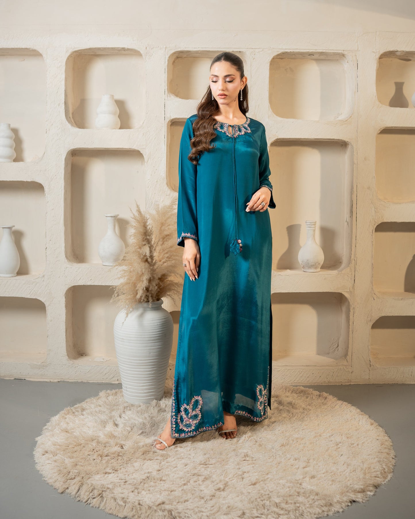 Woman in a teal dress standing in front of a decorative wall with shelves and vases.
