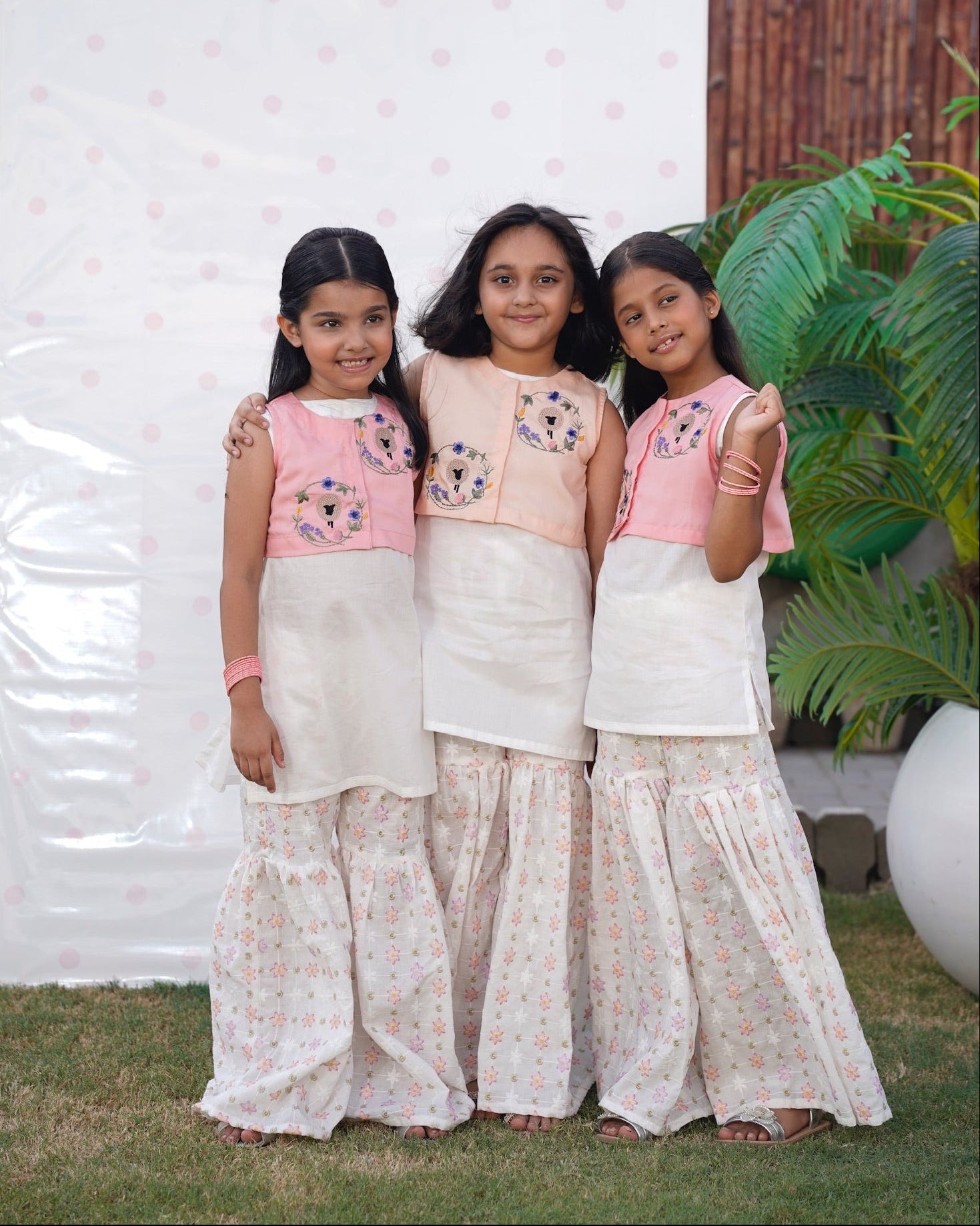 Three young girls in matching traditional outfits standing outdoors with a white wall and plant in the background. Full front, standing outdoors: "Blush pink embroidered cotton koti with floral chikankari gharara, full front view SHAFAÉ Goatie Blush"