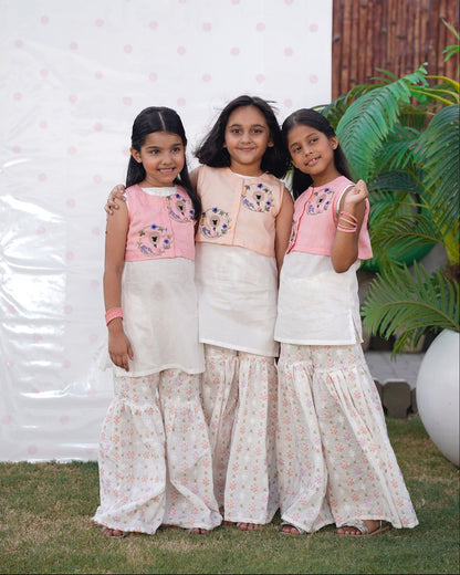 Three young girls in matching traditional outfits standing outdoors with a white wall and plant in the background. Full front, standing outdoors: "Blush pink embroidered cotton koti with floral chikankari gharara, full front view SHAFAÉ Goatie Blush"