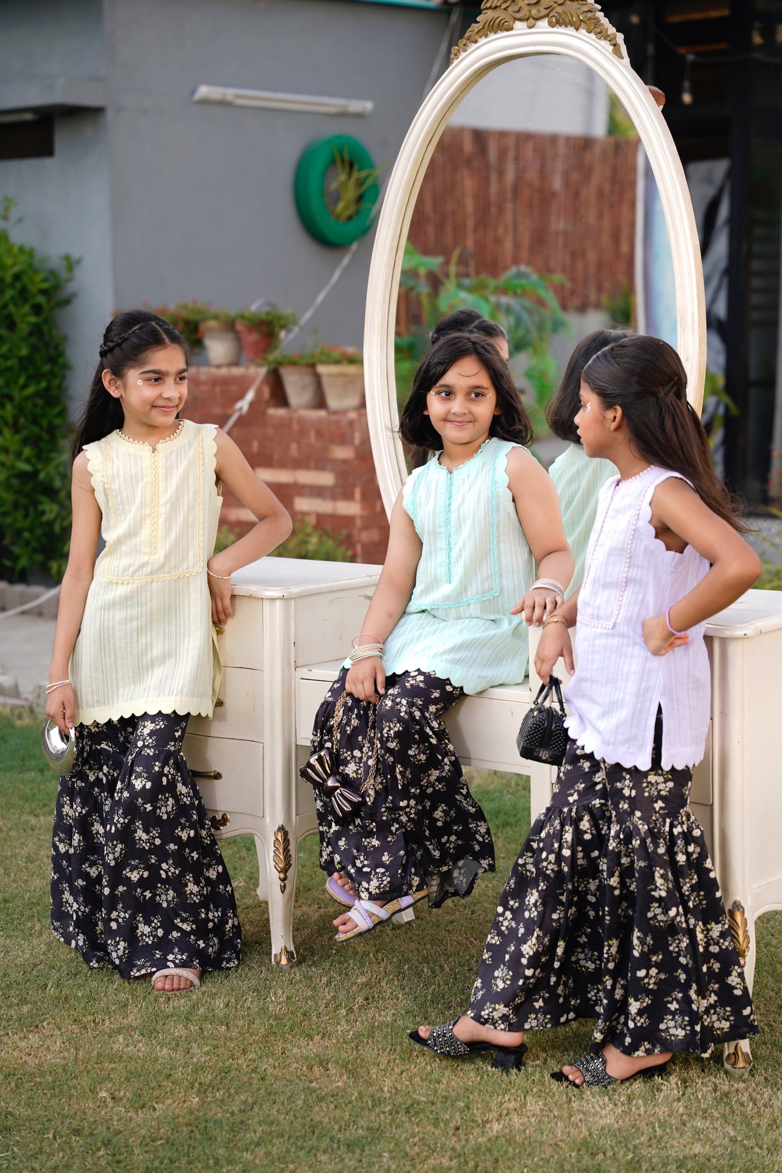 Three young girls in traditional outfits standing in front of a mirror outdoors.