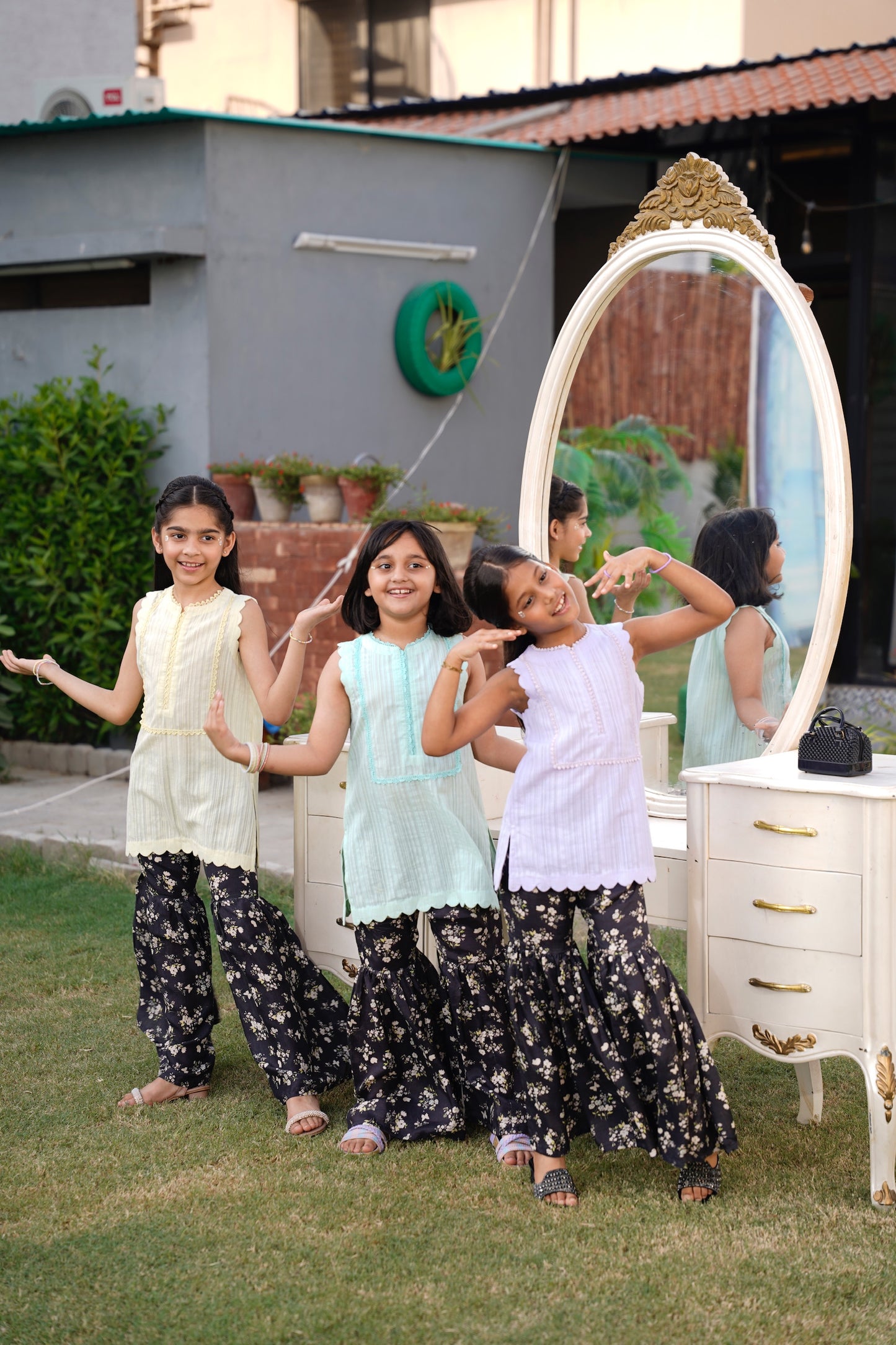 three young girls posing in front of a large decorative mirror outdoors. Full front, standing outdoors: "Lemon lawn sleeveless shirt with dark floral printed gharara, full front view SHAFAÉ Lemon Bloom"