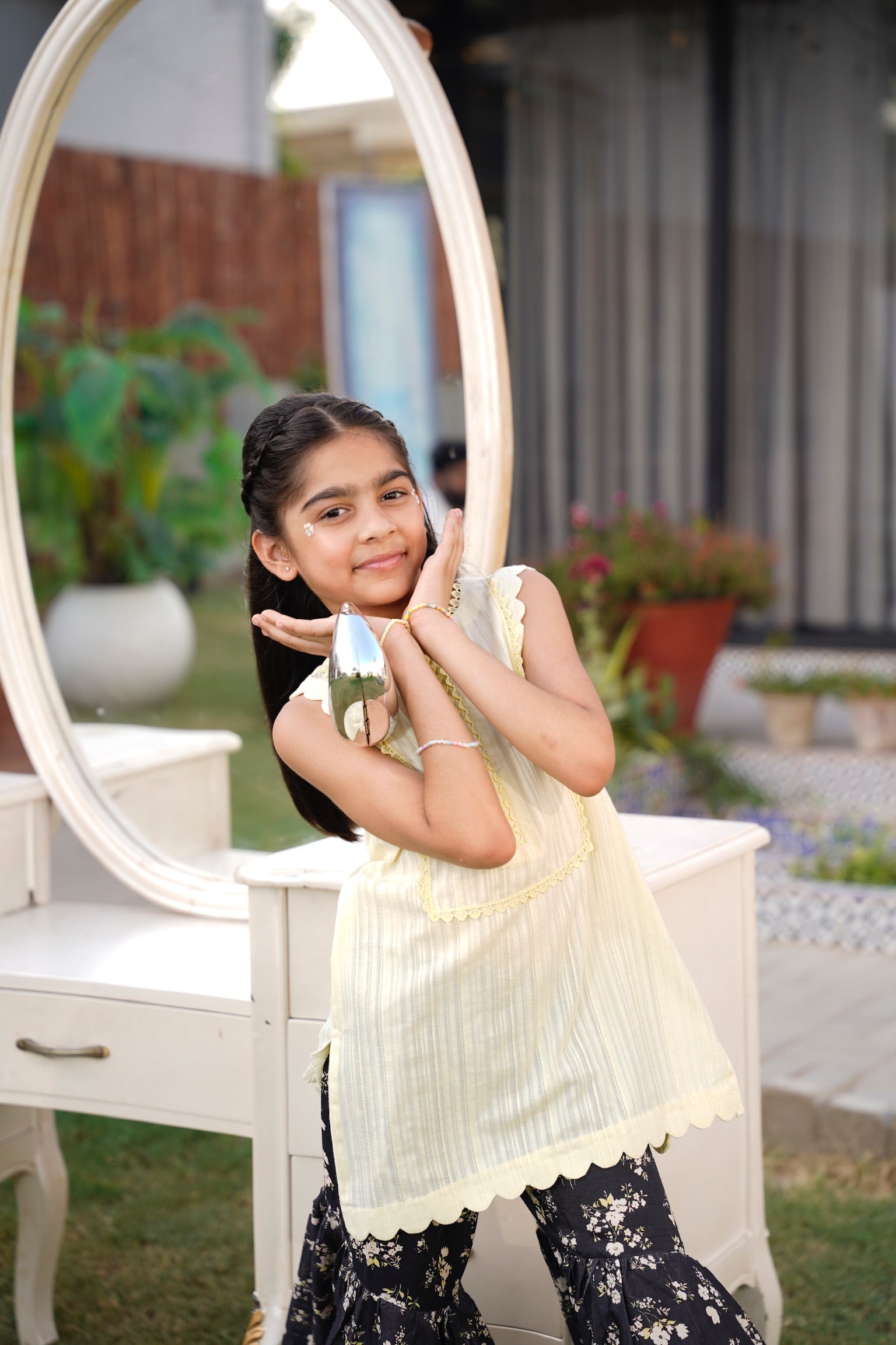 Young girl standing in front of a mirror outdoors, holding a small object.