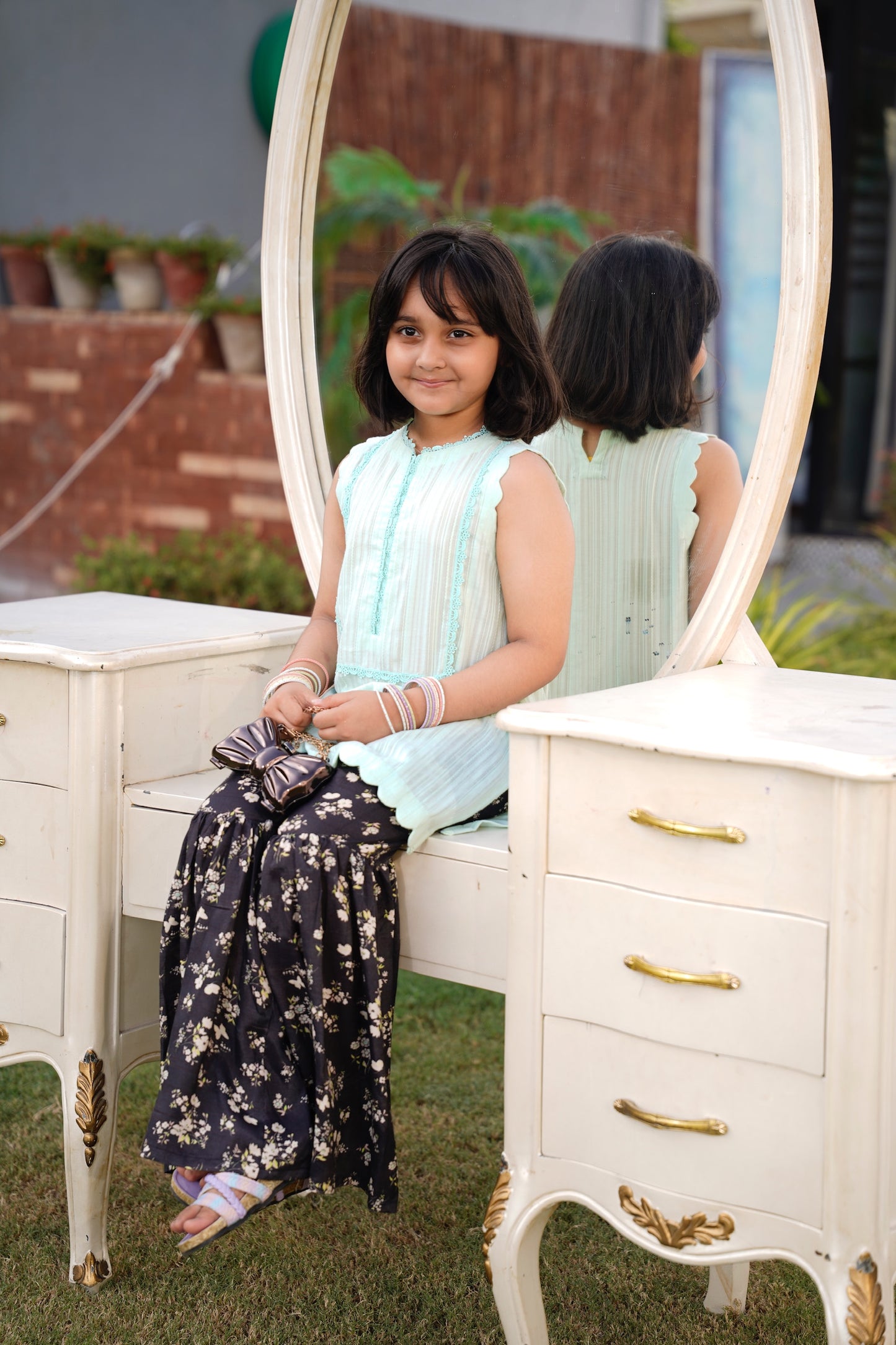 a young girls sitting on a white vanity mirror with a garden background Full front, standing outdoors: "Mint lawn sleeveless shirt with dark floral printed gharara, SHAFAÉ Mint Bloom"