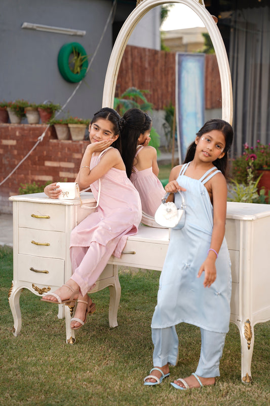 Two young girls in formal attire sitting on a white vanity table outdoors.  