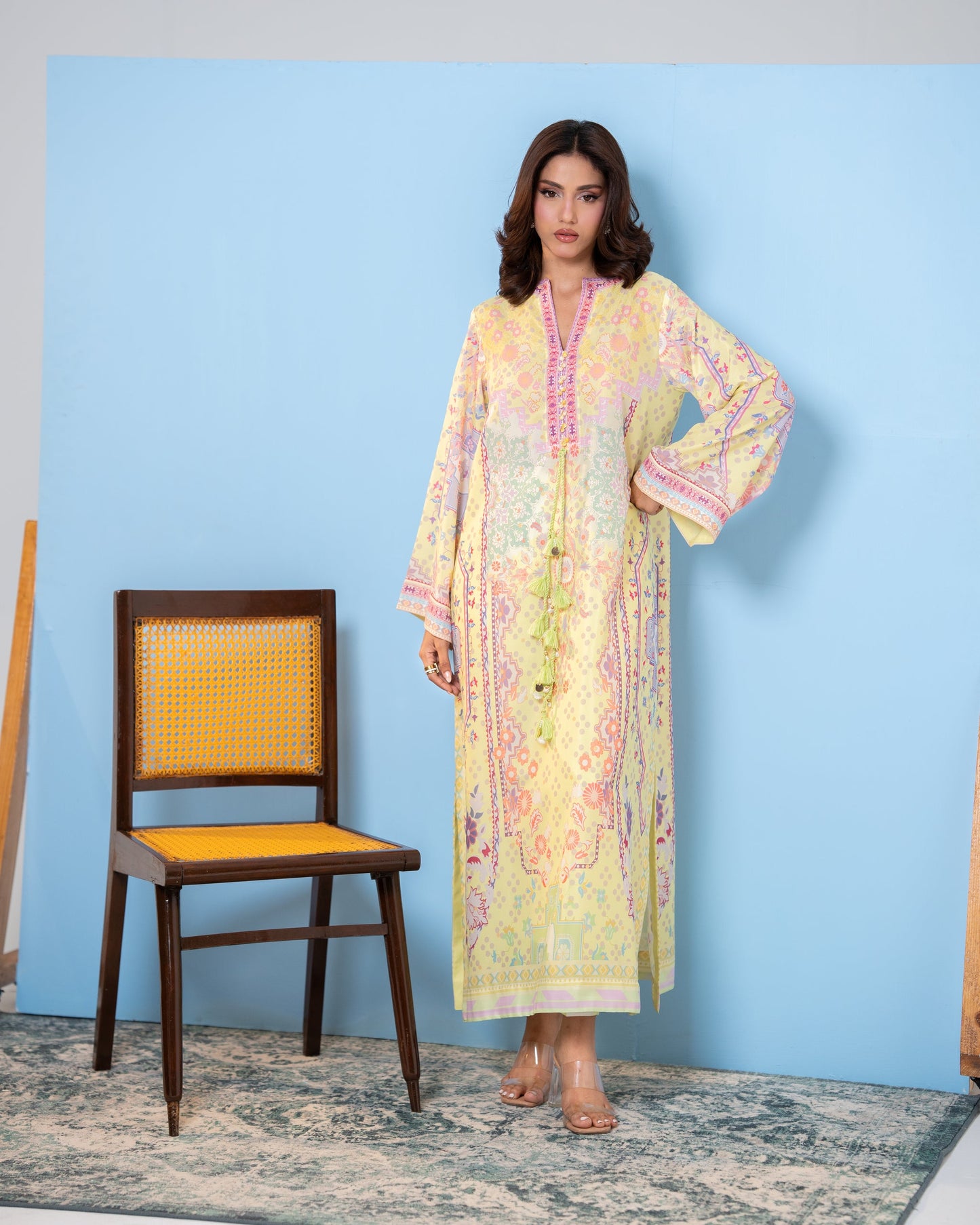 Woman in a pastel floral dress standing next to a chair against a blue wall. Pastel yellow printed silk kaftan, full front view SHAFAÉ Sunlit Kaftan"