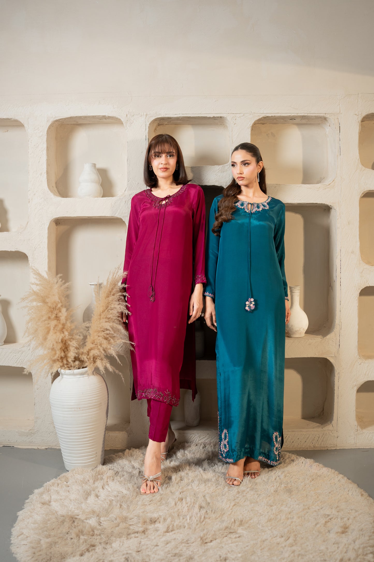 Two women in colorful outfits standing against a textured wall with decorative elements.