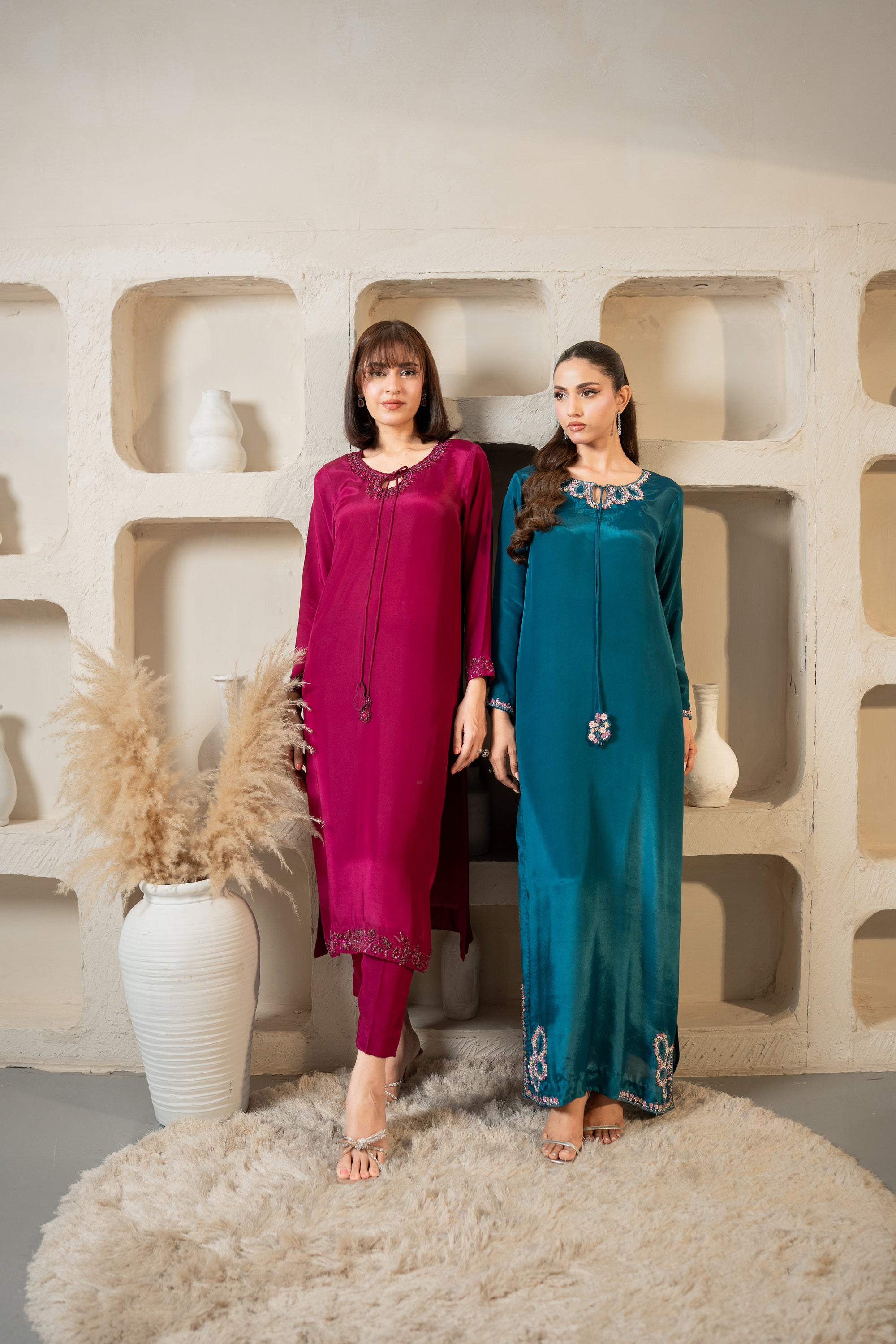 Two women in colorful outfits standing against a textured wall with decorative elements.