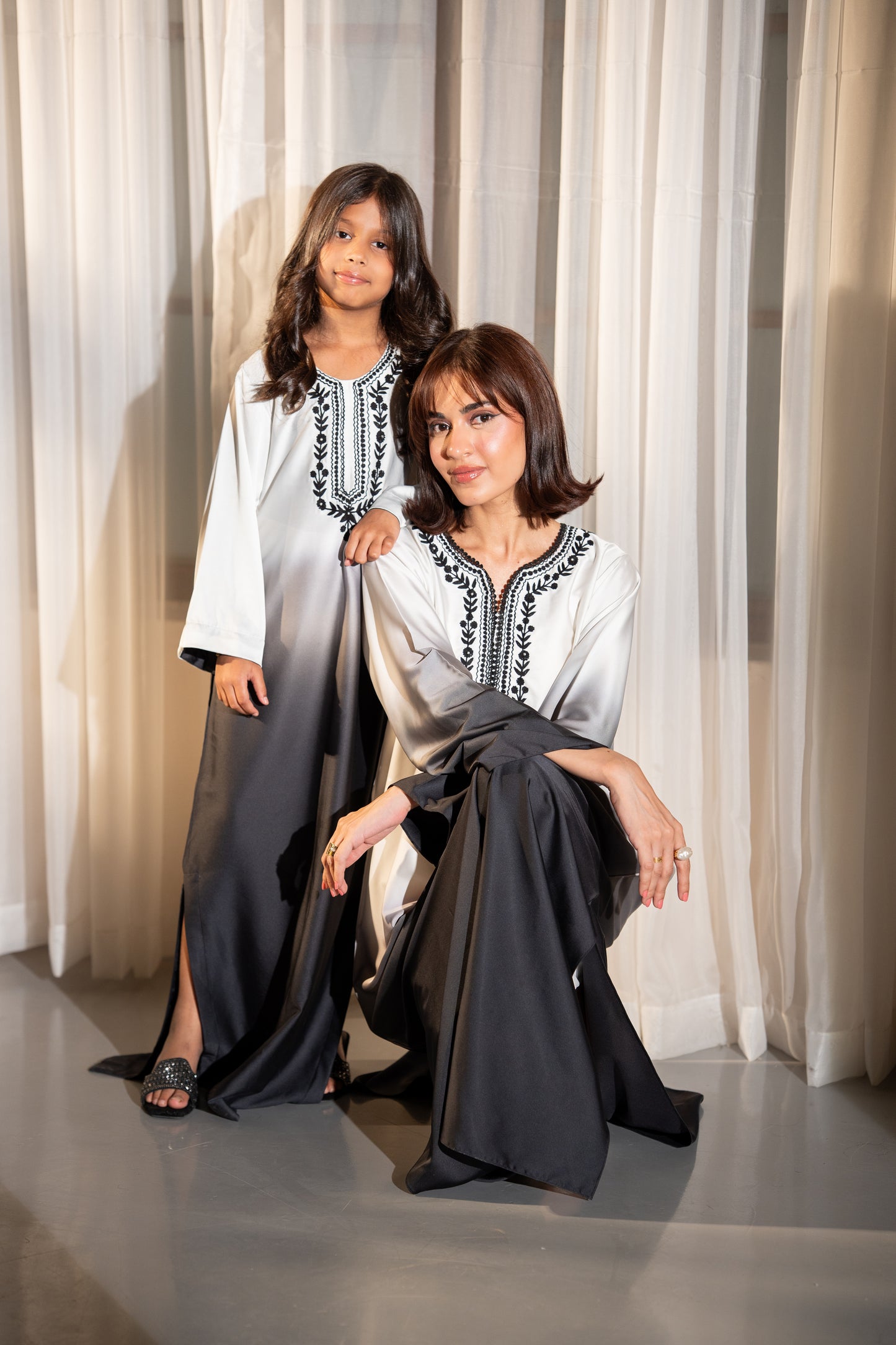 Two women in matching outfits posing against a white curtain. "Ombré silk kaftan fading from ivory to black, SHAFAÉ Noir Ombre Kaftan"